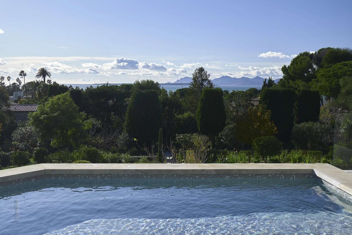 A serene infinity pool view of a lush green landscape, sparkling blue sea, distant mountains, and a bright sky.