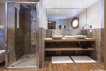 Modern bathroom with a glass shower, double vessel sinks on a wooden vanity, large mirror, and a reflected porthole window.