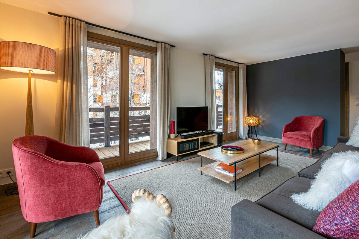Cozy living room with red armchairs, a gray sofa, TV, wooden coffee table, and large balcony doors showing an outdoor view.