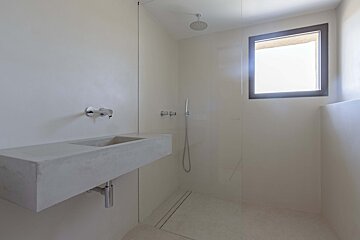 Bright, minimalist bathroom with a floating concrete sink, walk-in shower, and window, all in light tones.
