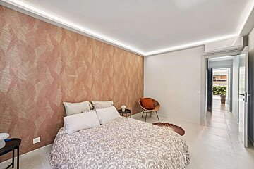 Modern bedroom with a salmon patterned accent wall, white bed, wicker chair, and hidden ceiling lights. A doorway leads to an open hallway.