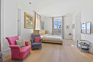 A chic, bright bedroom with white walls, light wood floor, a simple bed, vibrant armchairs, colorful art, and a large window with a city view.