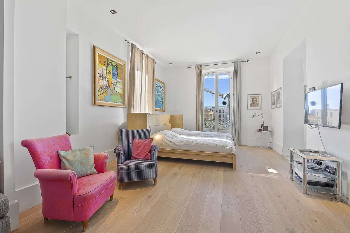 Bright bedroom with a light wood floor, a bed, pink and striped armchairs, a wall-mounted TV, and a large window overlooking a city.
