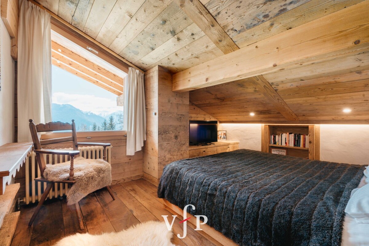 Cozy rustic chalet bedroom with sloped wooden ceilings, a plush bed, and a wooden chair by a window with mountain views.