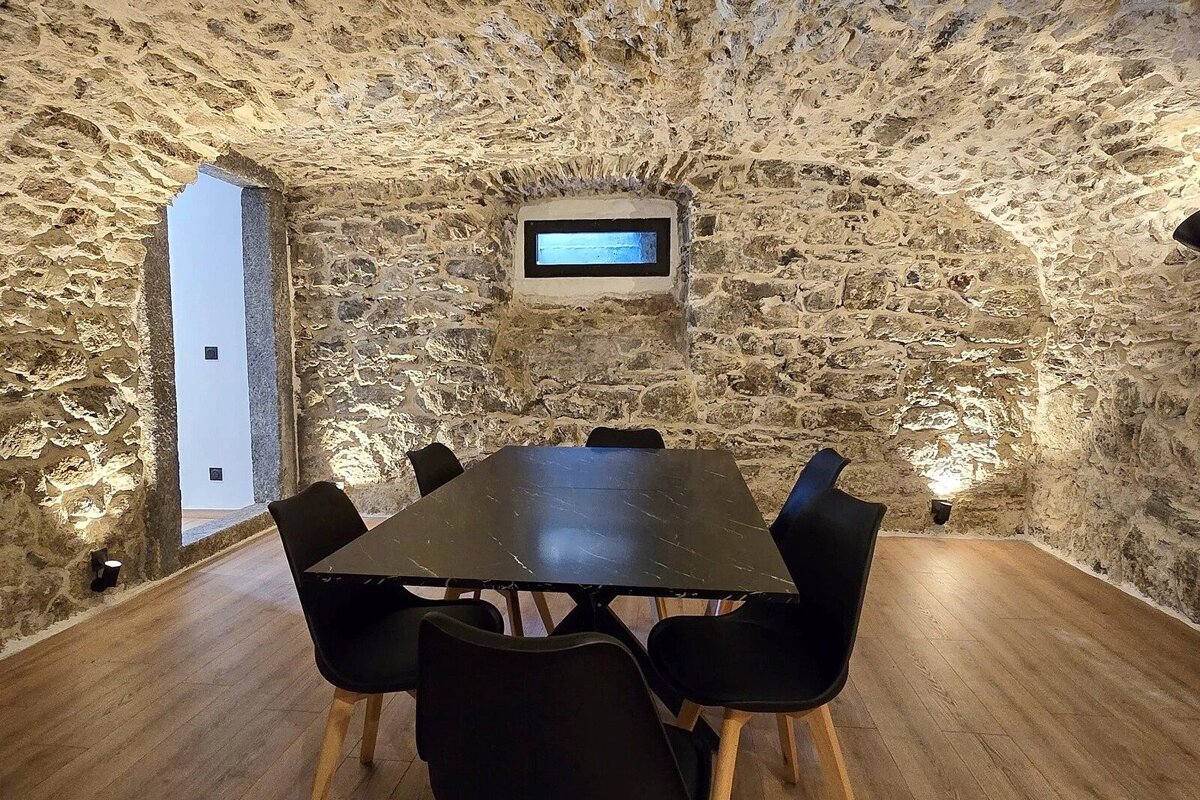 A cozy, stone-walled cellar dining room with an arched ceiling, modern black table and chairs, and warm accent lighting on a wooden floor.