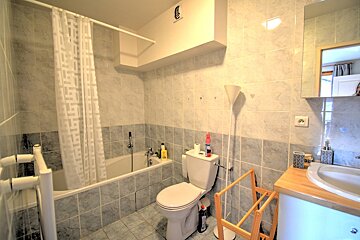 A tiled bathroom features a bathtub with a shower curtain, a toilet, a sink with a mirror above it, and a modern white floor lamp.