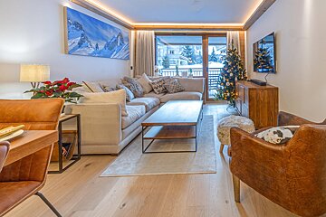 A warm, modern chalet living room with a large sofa, coffee table, Christmas tree, mountain art, and a balcony with snowy views.