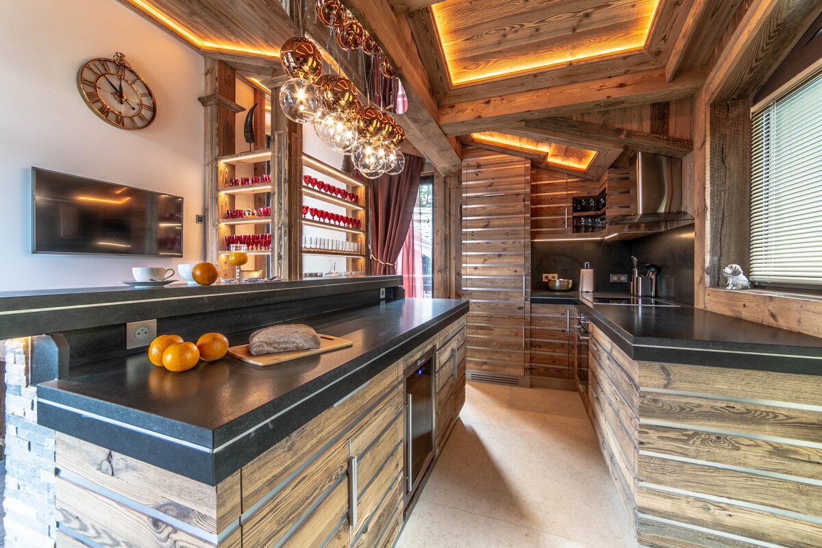A luxurious rustic kitchen/bar area featuring warm wooden beams, modern lighting, dark countertops, a TV, and fresh fruit.