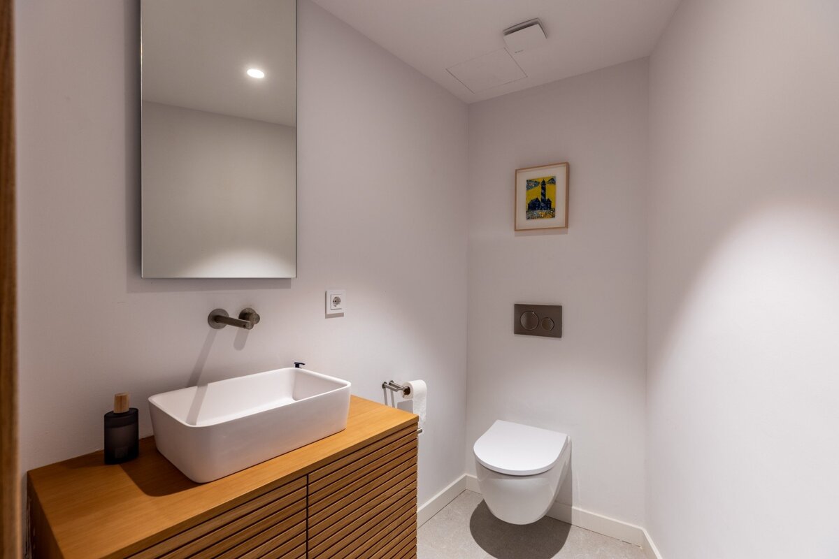 Clean, modern white bathroom featuring a wooden vanity, rectangular sink, wall-mounted toilet, mirror, and framed art.