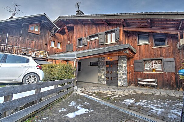 Wooden chalet-style house with a stone-paved driveway, a white car parked outside, and light snow on the ground.