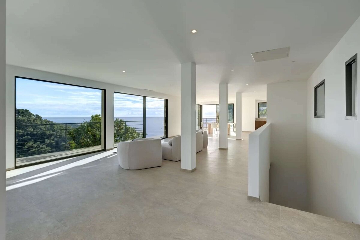 A bright, minimalist room with floor-to-ceiling windows offers a stunning panoramic view of the ocean and lush trees. White seating and light floors complete the modern aesthetic.