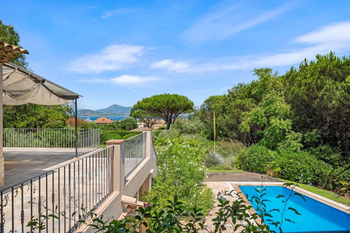 A stunning terrace view with a bright blue sky, distant sea, lush green trees, and a sparkling swimming pool below.