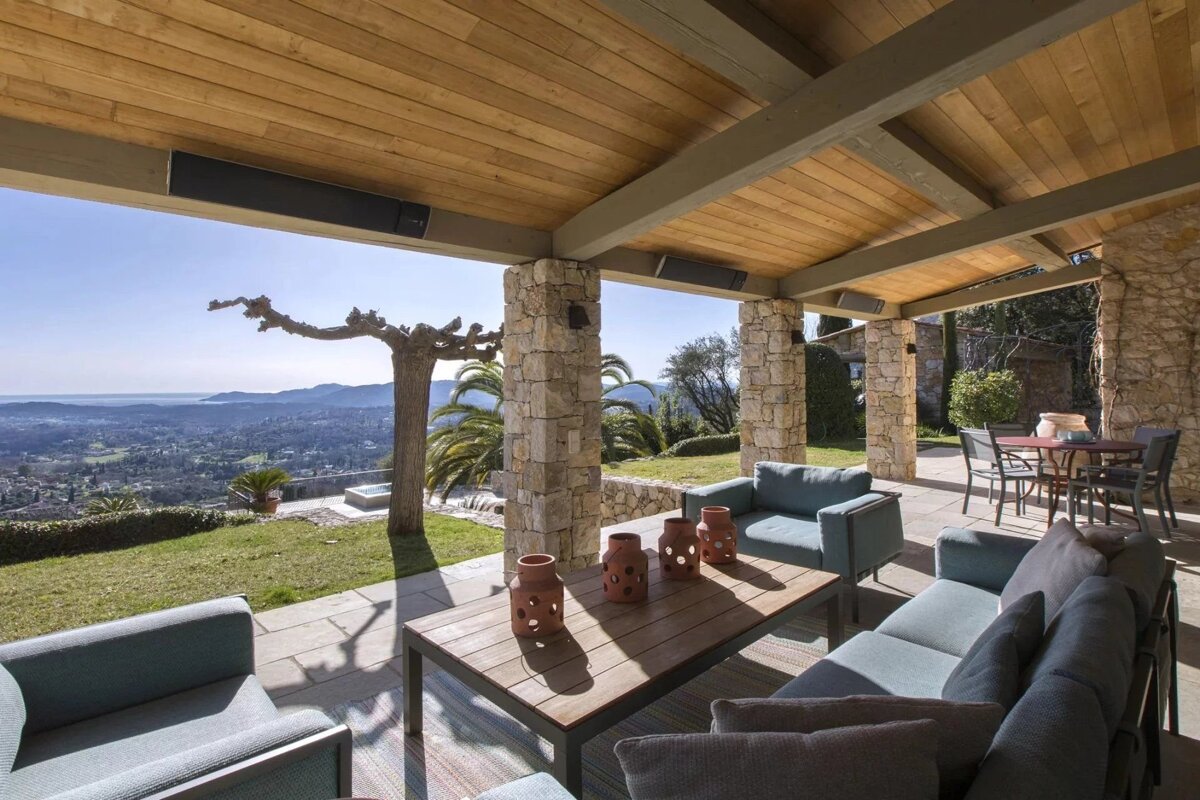 Elegant covered patio featuring teal sofas, stone columns, wooden ceiling, and heaters, overlooking a scenic valley & distant coastline.