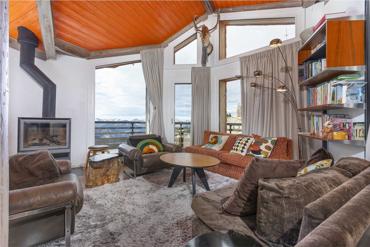 A warm, rustic chalet living room with a vibrant orange ceiling, cozy fireplace, comfy seating, and stunning panoramic mountain views.