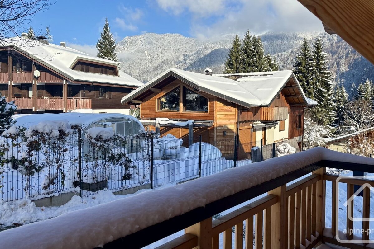 Snowy view of wooden mountain chalets and pine forests under a blue sky, shot from a snow-covered balcony.