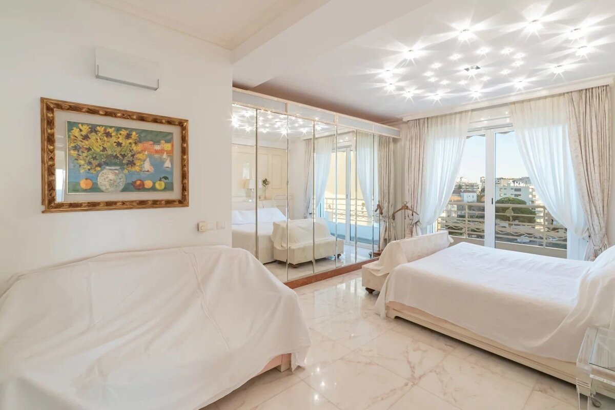A bright white bedroom features a mirrored wall, a bed, a covered sofa, and a colorful painting. A balcony offers a city view.