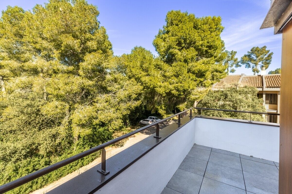 A modern balcony with a tiled floor and dark railing offers a view of lush green trees, a bright blue sky, and a distant street with parked cars.