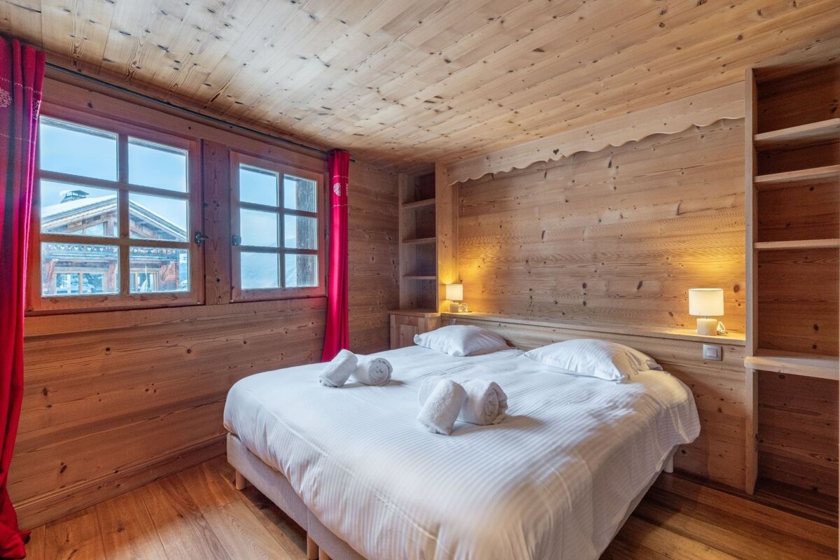 A cozy, rustic wooden bedroom in a chalet with a double bed, red curtains, warm lighting, and views of snowy buildings.