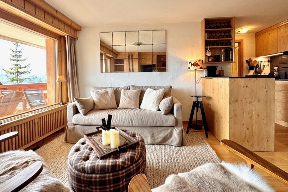 Cozy cabin living room with a beige sofa, plaid ottoman, grid-patterned mirror, and large window overlooking snowy trees.