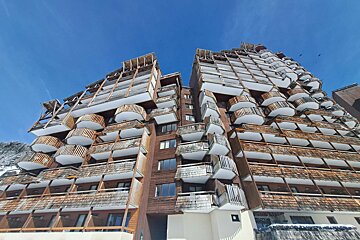 A towering brown wooden building with numerous curved balconies, resembling a mountain ski resort, against a clear blue sky.