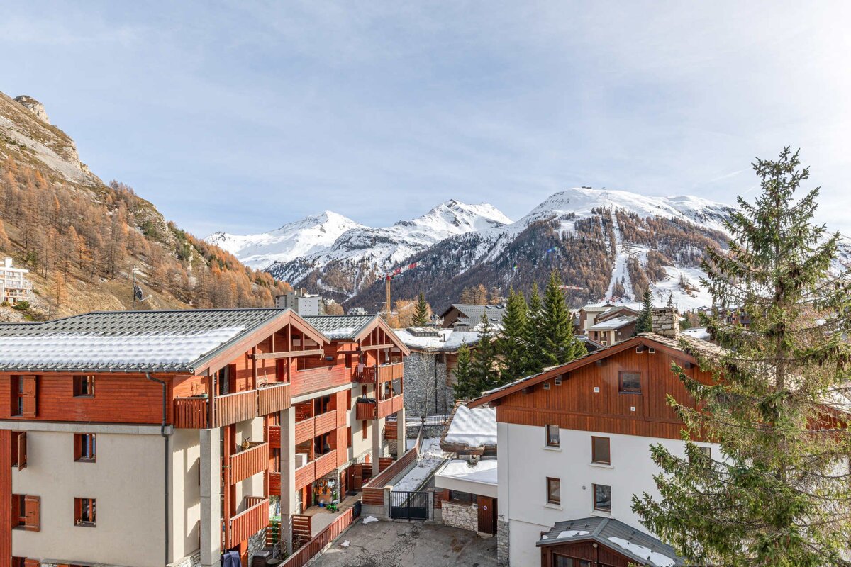 Panoramic view of a winter alpine village with wooden buildings and evergreen trees, framed by snow-covered mountains and ski slopes.