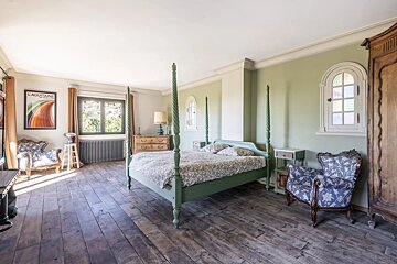 A bedroom with sage green walls, a four-poster bed, and blue patterned armchairs on hardwood floors.