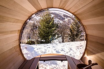 A cozy wooden barrel sauna's circular window frames a picturesque snowy mountain landscape with pine trees, offering a warm view of winter.
