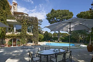 Sunny villa patio with outdoor dining, a swimming pool, and a large umbrella, beside a vine-covered house under blue skies.