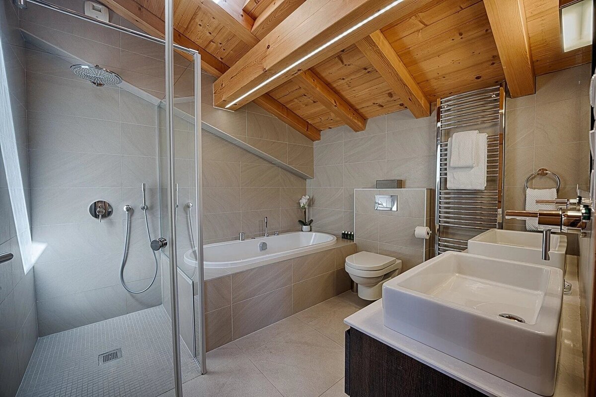A modern, spacious bathroom features a walk-in shower, bathtub, toilet, double vessel sinks, and a wooden beamed ceiling with integrated lighting.