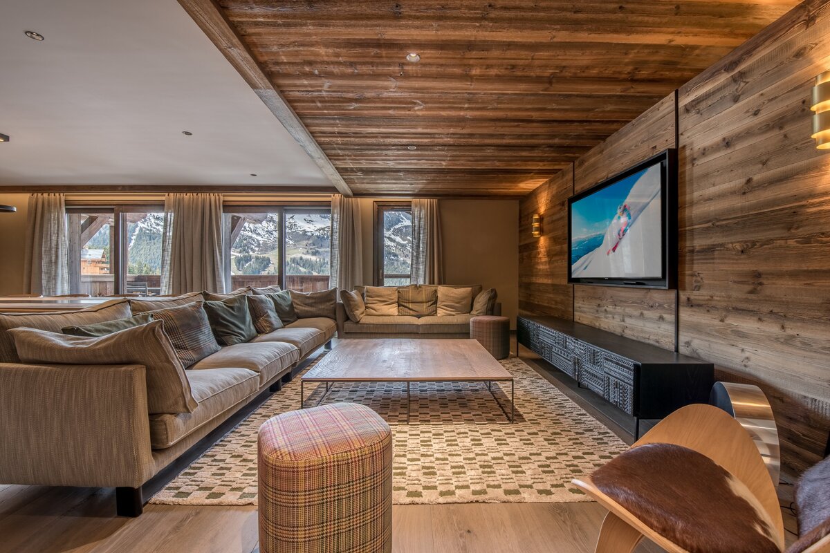 A rustic modern living room with extensive wood paneling, comfy sofas, a large TV, and a beautiful mountain vista outside.