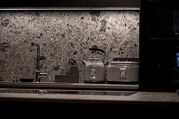 Modern kitchen counter with a grey terrazzo-patterned backsplash, under-cabinet lighting, a faucet, and matching grey kettle and toaster.