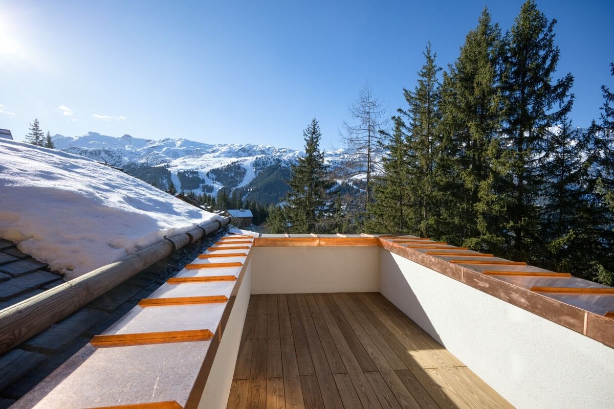 A sunny balcony with wooden floor and copper trim overlooks snowy mountains, pine trees, and ski slopes. A snow-covered roof is in the foreground.