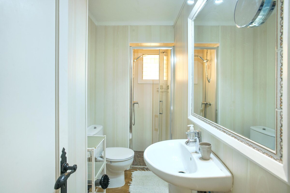 A small, brightly lit bathroom with a white pedestal sink, toilet, glass shower, and a large framed mirror. It features light-colored, striped walls.