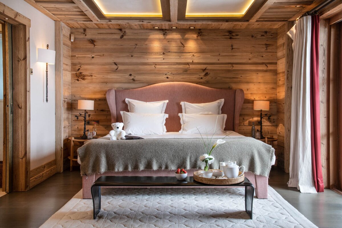 A cozy, rustic bedroom with wooden walls/ceiling, featuring a pink upholstered bed, white linens, and warm ambient lighting. A tray with tea and strawberries sits on a bench.