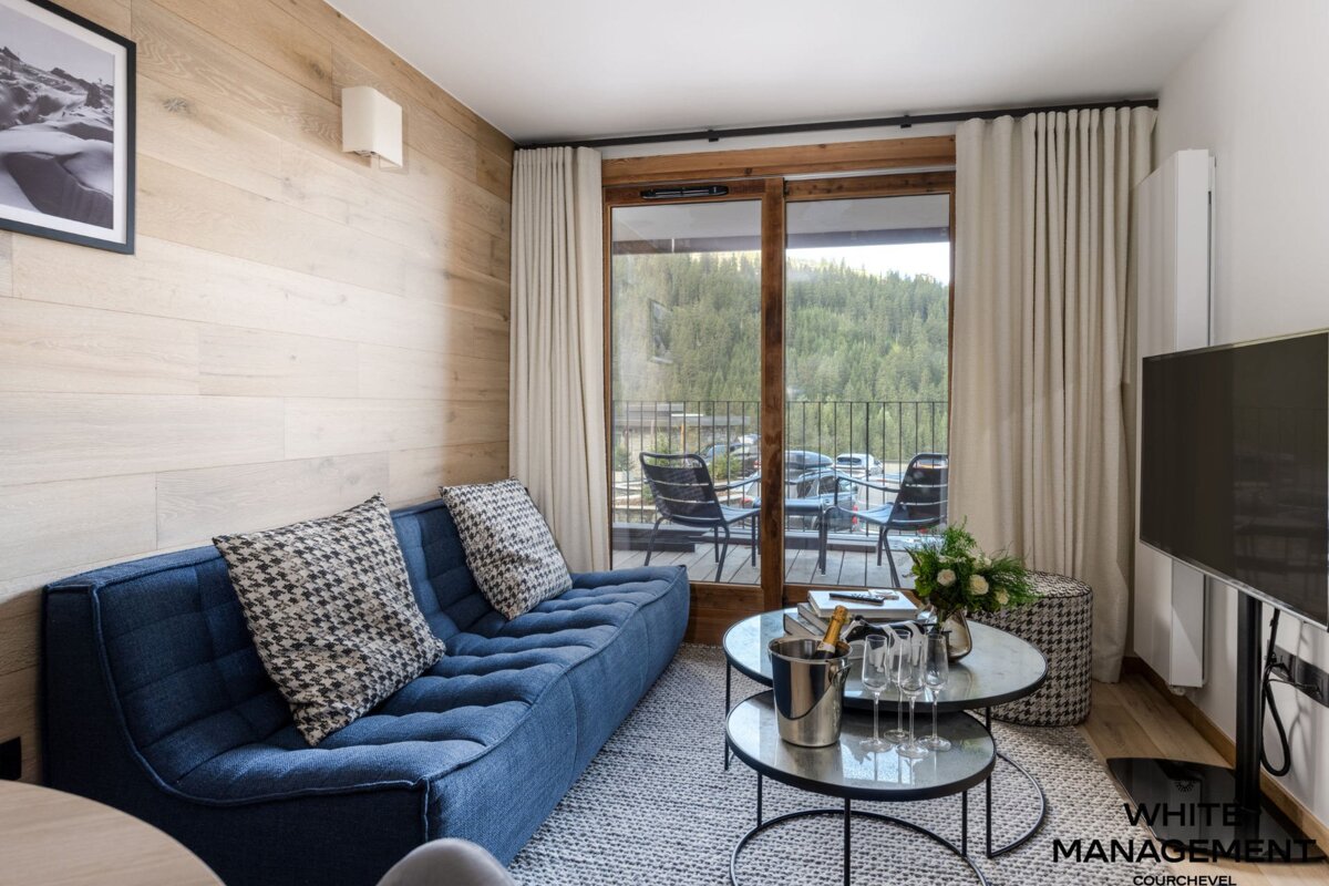 Cozy living room with a blue sofa, wood-paneled wall, and a balcony overlooking a lush forest. Champagne bucket on the coffee table.