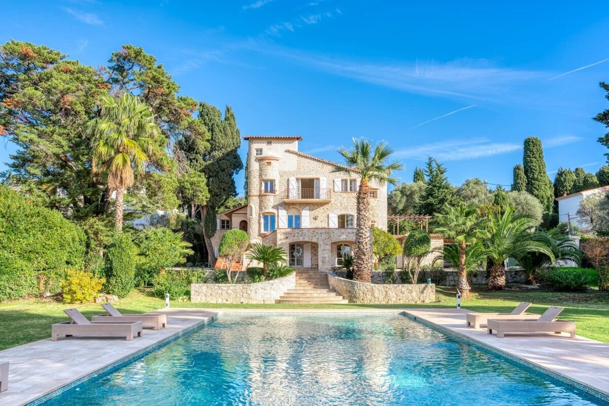 A large stone villa with a swimming pool, sun loungers, palm trees, and lush greenery under a clear blue sky.