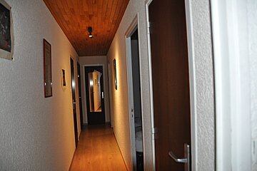 A narrow hallway features a wooden floor and ceiling, textured white walls with art, and multiple doors. Warm lighting creates shadows, and a mirror reflects further down the hall.