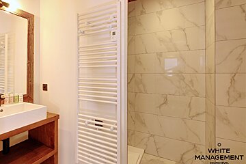 A modern bathroom featuring a wooden-framed mirror, sink on a wooden vanity, a white towel warmer, and a shower with light marble-look tiled walls.