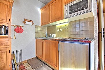 A cozy, compact kitchen with light wood cabinets, a sink, stove, microwave, and kettle. Red pot holders hang on the wall.