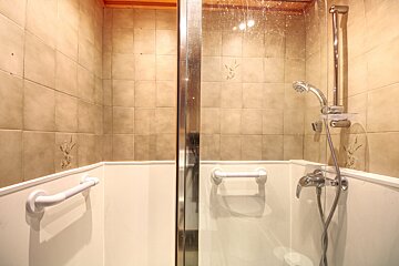 A shower cabin with light brown tiled walls, white lower panels, grab bars, and a shower head, enclosed by a glass panel.