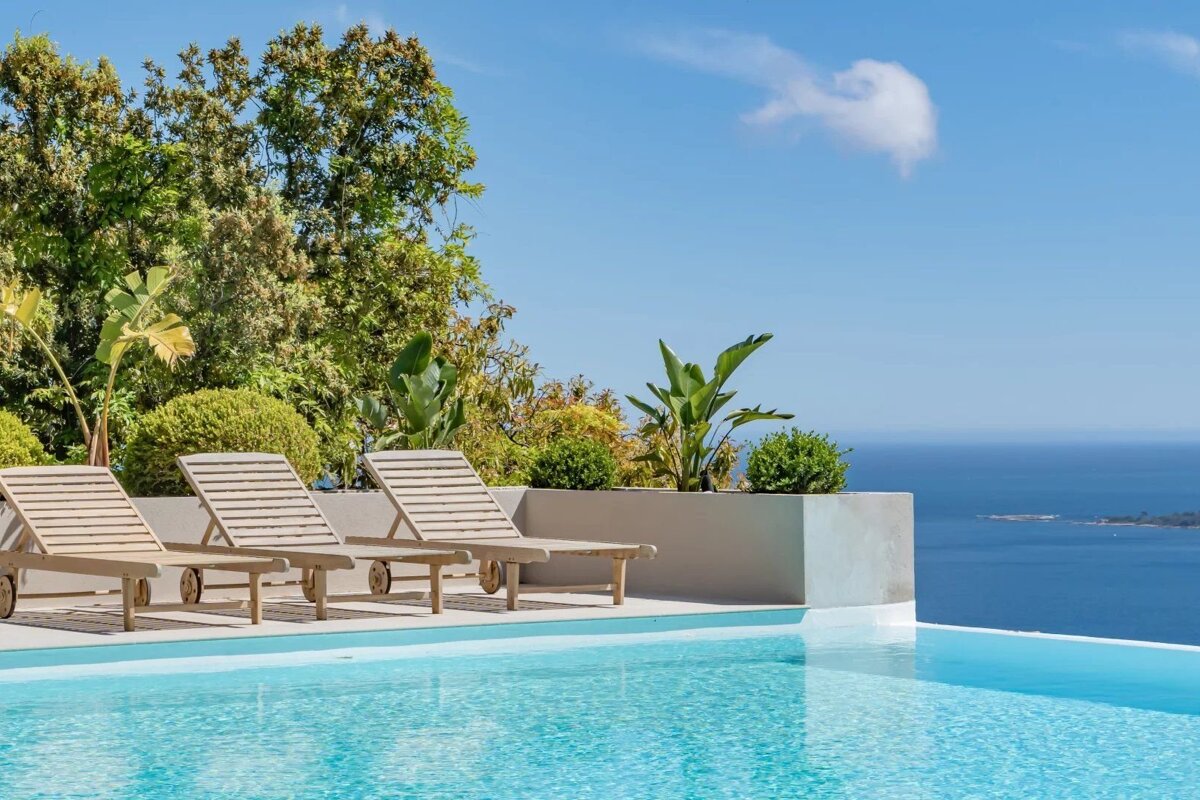 Three wooden lounge chairs sit by a stunning infinity pool overlooking the calm blue ocean under a clear sky.