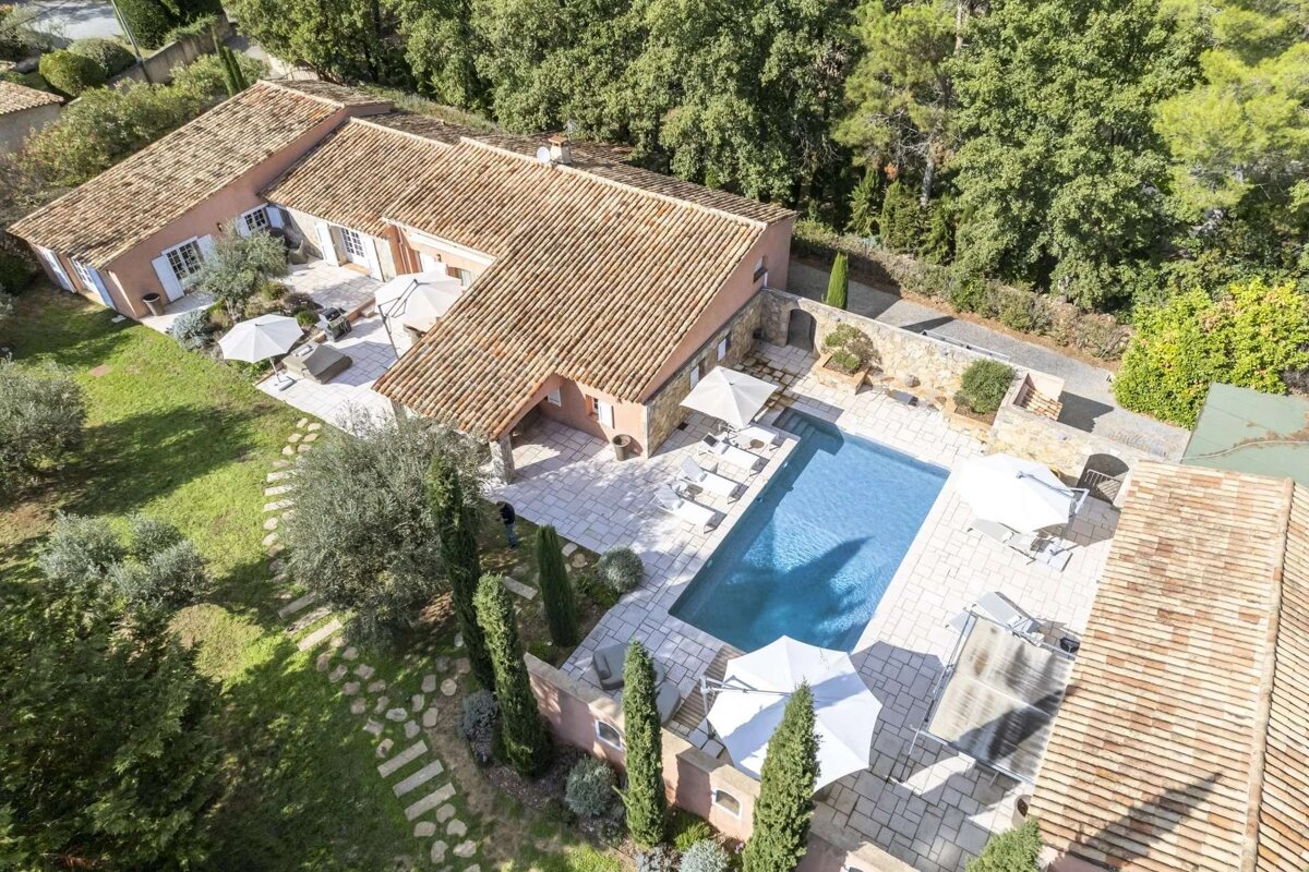 Aerial view of a luxurious villa with terracotta roofs, multiple patios, and a blue swimming pool, surrounded by lush green trees and gardens.