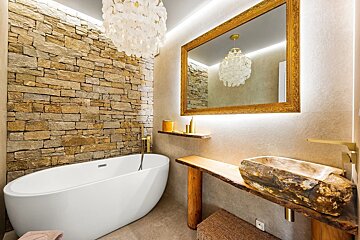 A rustic yet luxurious bathroom with a stone accent wall, white freestanding tub, a unique stone sink, and a grand ornate mirror.