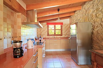A rustic-style kitchen with stone walls, wooden beams, tiled floors, and modern appliances. It features wood cabinets, red countertops, and a window.