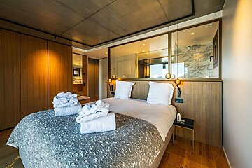 A modern bedroom with a large bed, grey throw, and white robes. It features wood paneling, a concrete ceiling, and a mirrored wall to the bathroom.