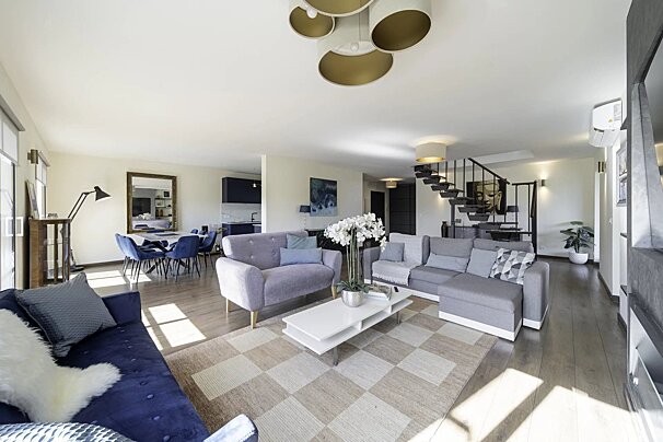 An open-plan living room with grey and blue sofas, a dining area, wooden floors, and a modern open staircase.