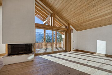 Empty chalet room with a fireplace, vaulted wood ceiling, and sliding glass doors opening to snowy mountain views.