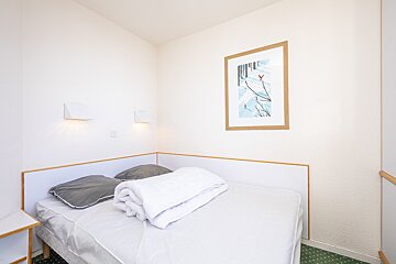 A minimalist bedroom with a single bed, white bedding, and grey pillows against white walls. A framed winter scene art and lamps are visible. Green carpet.