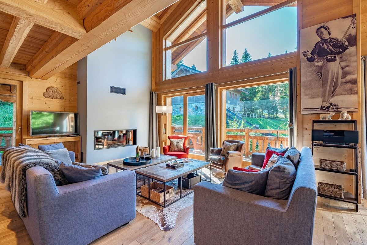 Spacious, warm wooden living room with a modern fireplace, comfy sofas, and large windows offering a lush green mountain view. A large ski poster adorns a wall.