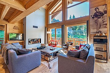 Spacious, warm wooden living room with a modern fireplace, comfy sofas, and large windows offering a lush green mountain view. A large ski poster adorns a wall.